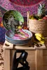 A collection of decorative plates with animal and floral designs, a basket of fruit, and a sculpture are arranged on a wooden surface against a colorful tapestry background. Anteprima dell’immagine prodotto
