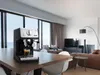 An espresso machine sits on a wooden table in a modern living room with large windows overlooking the sea. Voir les images du produit