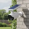 A solar-powered security camera is mounted on a brick wall, with greenery in the foreground and background. Produktbild-Vorschau