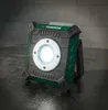 A portable LED work light is placed on a dark, textured surface in front of a brick wall. Produktbild-Vorschau