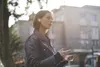 A woman wearing earbuds and holding a phone, standing outdoors with buildings in the background. Anteprima dell’immagine prodotto