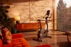 An exercise bike is placed in a living room with a sofa, a cabinet, and a large window overlooking a natural landscape. Produktbild-Vorschau