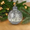 A decorative Christmas ornament with a winter forest scene and snowflakes, placed on a wooden surface with a blurred Christmas tree and lights in the background. Produktbild-Vorschau