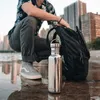 A person crouches next to a backpack and a stainless steel water bottle on a wet surface with city buildings in the background. Podgląd zdjęcia produktu