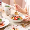 A person is serving a fruit salad from a bowl with a golden spoon, with other tableware visible on a table. Voir les images du produit