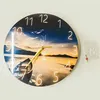 A round wall clock with a sunset landscape image is mounted on a white wall. A small bag with mounting hardware is next to the clock. Voir les images du produit