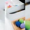 A close-up shot of a white curved siphon cover, with clear plastic containers holding colorful hair accessories in the background, and a wooden tray with colorful balls in the foreground. Produktbild-Vorschau