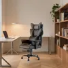 A black ergonomic office chair with a footrest is placed in a home office setting next to a desk with a laptop and a lamp. A bookshelf filled with books is visible in the background. Produktbild-Vorschau