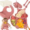 A baby is eating from a silicone feeding set that includes a bib, plate, cup, and utensils. The set is pink and shaped like a squirrel. Voir les images du produit