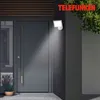 An outdoor LED light with a motion sensor is mounted on a dark gray wall next to a modern gray door. The light illuminates the door and the surrounding area. A potted plant with lavender is visible near the door. Produktbild-Vorschau