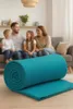 A family is sitting on a couch in a living room, with a rolled-up teal blanket in the foreground. Produktbild-Vorschau