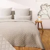 A neatly made bed with a beige quilted bedspread and pillows, in a room with a pink wall and a wooden stool with a lamp. Anteprima dell’immagine prodotto