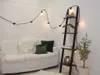 A cozy living room corner with a white sofa, decorative pillows, a ladder shelf, and string lights. Produktbild-Vorschau
