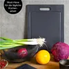 A cutting board with various fresh vegetables and fruits arranged in front of it on a wooden surface. Anteprima dell’immagine prodotto