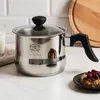 A stainless steel milk pot with a glass lid and black handle sits on a marble countertop, next to a glass of milk and a small plate with food. Náhled obrázku produktu