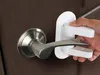 A hand is shown attaching a white safety lock to a silver door handle on a brown door. Náhled obrázku produktu