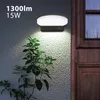 An outdoor LED lamp is mounted on a textured wall next to a wooden door, with a plant in the foreground. Podgląd zdjęcia produktu