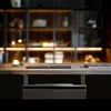 A close-up shot of a desk with a laptop and a drawer underneath. The background shows a blurred bookshelf with lights. Voir les images du produit