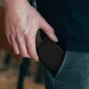 A person's hand is shown inserting a black object into their pants pocket. Produktbild-Vorschau