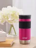 A pink Contigo travel mug sits on a wooden surface next to a white gift box and a stack of books, with a vase of white flowers in the background. Anteprima dell’immagine prodotto