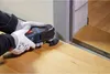 A person wearing gloves is using an oscillating multi-tool to cut along the edge of a wooden floor next to a doorway. Podgląd zdjęcia produktu