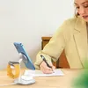 A woman is writing on a piece of paper at a desk, with a 3-in-1 wireless charging station for a phone, smartwatch, and earbuds in the foreground. Produktbild-Vorschau