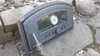 A cast iron pizza oven door with a glass window and thermometer, resting on a stone patio. Produktbild-Vorschau