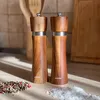 Two wooden salt and pepper grinders on a countertop with scattered peppercorns and salt. Náhled obrázku produktu