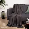 A dark gray fleece blanket is draped over a gray sofa, with a decorative black lantern containing artificial plants to the left and a small table with a teapot and cup to the right. Produktbild-Vorschau
