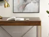 A close-up view of a wooden desk with an open book and a small golden object on top, set against a white wall with abstract art above. Voir les images du produit