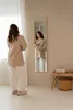 A woman stands in front of a full-length mirror, looking at her reflection. The room has a minimalist aesthetic with neutral tones. Náhled obrázku produktu