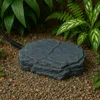 A rock-shaped heating element is placed on gravel, surrounded by lush green plants. Anteprima dell’immagine prodotto