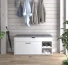 A shoe bench with a padded seat and storage compartments, set against a wood-paneled wall with coats and hats hanging above. An umbrella is leaning against the wall next to the bench. A plant is visible on the right side. Voir les images du produit
