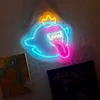 A neon sign of a ghost character with a crown and tongue sticking out, hanging on a textured wall next to two framed pictures. Voir les images du produit