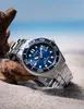 A watch is placed on sand next to a piece of driftwood, with a blurred beach and ocean background. Anteprima dell’immagine prodotto