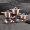 A set of rose gold cookware is displayed on a dark countertop against a textured black stone wall. Podgląd zdjęcia produktu