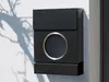 A black mailbox mounted on a white wall with tree shadows. Produktbild-Vorschau