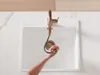 A person's hand is using a copper kitchen faucet with a pull-out spray in a white sink. Produktbild-Vorschau