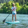 A woman is paddleboarding on a lake with trees in the background. Produktbild-Vorschau