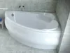 A white corner bathtub with a curved front panel is shown in a bathroom with gray tiled walls and floor. Produktbild-Vorschau