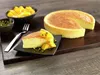 A cheesecake with mango topping and pistachios is presented on a wooden board. Produktbild-Vorschau