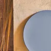 A close-up view of a round, gray-blue table with dried grass stalks on the left, against a textured beige wall. Anteprima dell’immagine prodotto