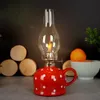 A red and white polka dot oil lamp with a lit flame sits on a gray surface, with wooden discs and a bowl of fruit in the background. Náhled obrázku produktu