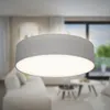 A close-up of a grey fabric drum pendant light with a white diffuser, set against a blurred background of a modern living room with furniture and a window. Podgląd zdjęcia produktu