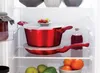 A set of red pots and pans with lids are neatly stacked on a shelf inside a refrigerator. A small plate of red berries sits on top of the larger pot. To the right, a small glass bottle with a cork stopper contains a dark red liquid. Below, a bowl holds ora Anteprima dell’immagine prodotto