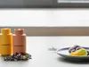 Two spice grinders are shown on a table next to some dried flowers, with a plate of food in the background. Produktbild-Vorschau