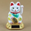 A white Maneki Neko (lucky cat) figurine with a red collar and green scarf, holding a gold coin with Chinese characters, sits on a gold base with a solar panel. The background is a plain light brown. Produktbild-Vorschau