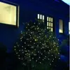 A bush decorated with LED lights at night, in front of a dark blue house with illuminated windows. Produktbild-Vorschau