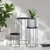 Three black metal plant stands of varying heights are arranged on a white surface with a marble-patterned wall in the background. A large green plant is on the left, and a smaller potted plant sits on the tallest stand on the right. A white candle is on th Náhľad obrázku produktu