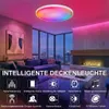 A modern living room with a large sectional sofa, a fireplace, and a large window overlooking a city skyline at night. The ceiling light is illuminated with colorful RGB lighting. Produktbild-Vorschau